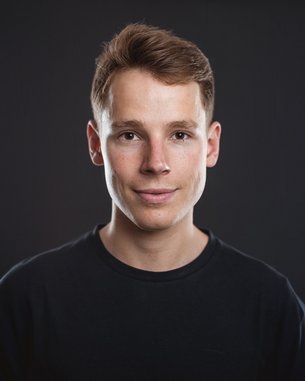 Ein junger Mann mit kurzem, braunem Haar und einem neutralen Gesichtsausdruck trägt ein schwarzes T-Shirt. Der Hintergrund ist dunkel und hebt seine Gesichtszüge hervor.