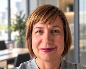 Frau mit schulterlangen, braunen Haaren und lila Lippenstift, lächelt in einem modernen Büro mit Pflanzen im Hintergrund.
