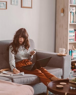 Eine Frau sitzt auf einem Sofa, nutzt einen Laptop und hält ein Smartphone in der Hand. Neben ihr liegen Unterlagen und eine Tasse auf einem Tisch. Im Hintergrund sind Bücherregale sichtbar.