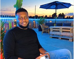 Ein Mann sitzt entspannt auf einer Terrasse, hält ein Smartphone in der Hand, umgeben von Pflanzen und einer gemütlichen Atmosphäre bei Sonnenuntergang.