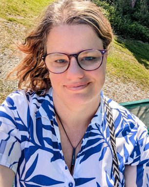 Eine Person mit Brille trägt ein blau-weißes Hemd und lächelt. Im Hintergrund ist eine grüne Landschaft zu sehen.
