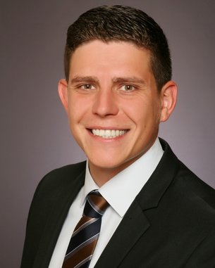 Ein junger Mann mit kurzen, dunklen Haaren und einem breiten Lächeln trägt einen schwarzen Anzug und eine gestreifte Krawatte. Der Hintergrund ist neutral und unauffällig.