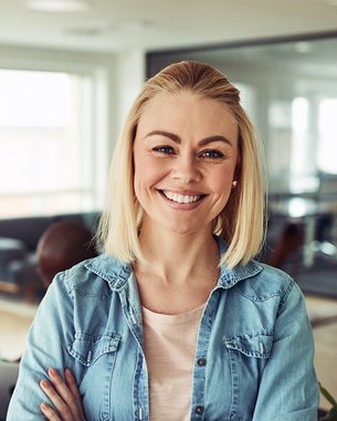 Eine lächelnde Frau mit blonden Haaren steht im Vordergrund eines modernen Büros, während im Hintergrund zwei Personen an einem Tisch arbeiten.