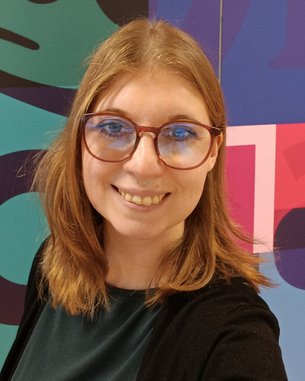 Eine Person mit langen, blonden Haaren und Brille lächelt in die Kamera. Im Hintergrund sind bunte geometrische Muster zu sehen.