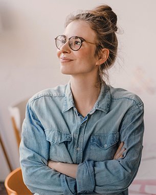 Junge Frau mit Brille und einem lässigen Jeanshemd, die mit verschränkten Armen nachdenklich schaut. Im Hintergrund sind Möbel und eine Lampe sichtbar.
